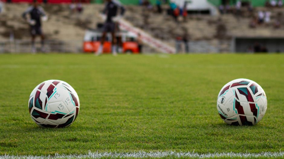 Two white soccer balls with black, red, and teal geometric patterns rest on a well-maintained green grass field, which appears freshly mowed and evenly trimmed. The turf surface looks smooth and clean, with visible white boundary lines at the front. In the background, blurred spectators sit in the stadium seating, and a portion of the stadium structure, including stairs and safety barriers, is visible under bright outdoor lighting. The scene suggests a clean, professional sports environment, consistent with surface cleaning and maintenance standards promoted by Carpet Cleaner Fulham, as part of their expertise in deep cleaning, sanitisation, and commercial cleaning for event venues.