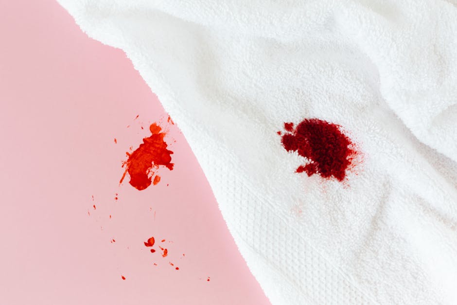 A white paper towel with a textured surface is shown on a pink background, stained with two red blood-like spots, one larger and more concentrated on the white towel, and a smaller, more dispersed stain on the pink surface. The scene illustrates an example of a fresh stain on household surfaces, emphasizing the importance of prompt stain removal in domestic cleaning. The image reflects the need for efficient surface cleaning and sanitisation, aligning with the focus of [PAGE_TITLE] on tackling common household stains. The cleanliness and condition of the surfaces highlight the potential for effective cleaning solutions offered by Carpet Cleaner Fulham, a specialist in deep cleaning and hygiene maintenance.