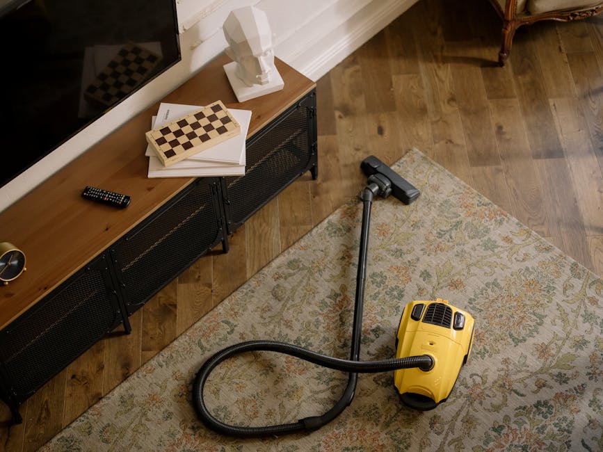 A yellow canister vacuum cleaner with a black hose and metal wand is positioned on a patterned area rug in a living room, ready for surface cleaning or deep cleaning. The room features a wooden floor, with a black metal and wood sideboard along the wall holding a remote control, a chessboard, papers, and a white bust sculpture. The space appears tidy and well-maintained, with natural light enhancing the clarity of the scene, illustrating professional domestic cleaning practices by Carpet Cleaner Fulham as part of their guide to carpet cleaning near Fulham Broadway SW6.