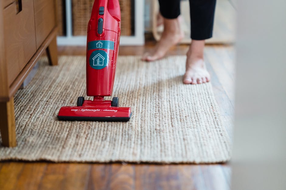 Close-up of a person vacuuming a beige and brown textured carpet using a red upright vacuum cleaner, with visible wheels and a power button. The person, wearing black leggings, is barefoot, and their feet are positioned on the carpet near a wooden cabinet. The room features wooden flooring adjacent to the carpet, with natural light illuminating the space. The vacuum cleaner shows branding and is positioned centrally, indicating a focus on surface cleaning and deep cleaning procedures. The environment appears clean and well-maintained, emphasizing professional domestic cleaning services offered by Carpet Cleaner Fulham as highlighted in the guide to carpet cleaning near Fulham Broadway SW6.