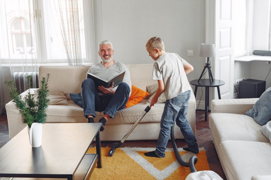 A yellow canister vacuum cleaner with a black hose and metal wand is positioned on a patterned area rug in a living room, ready for surface cleaning or deep cleaning. The room features a wooden floor, with a black metal and wood sideboard along the wall holding a remote control, a chessboard, papers, and a white bust sculpture. The space appears tidy and well-maintained, with natural light enhancing the clarity of the scene, illustrating professional domestic cleaning practices by Carpet Cleaner Fulham as part of their guide to carpet cleaning near Fulham Broadway SW6.
