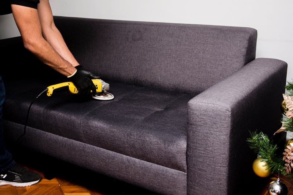 A person wearing black gloves is using a yellow handheld electric fabric steamer or upholstery cleaner on a dark gray fabric sofa in a living room. The sofa has a contemporary design with a high backrest and armrest, placed on a wooden floor. To the right of the sofa, there is a small decorated Christmas tree with gold and pink ornaments, indicating a seasonal setting. The room is brightly lit with natural light, highlighting the clean and maintained appearance of the sofa, which is being surface cleaned as part of deep upholstery sanitisation. The backdrop features a plain white wall, and the image is associated with residential cleaning services by Carpet Cleaner Fulham, focusing on upholstery cleaning tips for period flats.